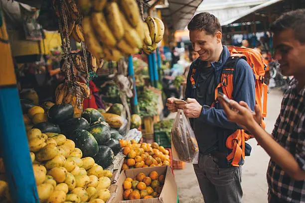 Photo of a tourist, on a local flea market, using a mobile phone while buying vegetables and fruit 