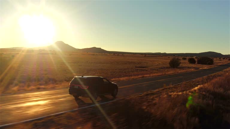 AERIAL: Black SUV car driving along empty country road at golden summer sunset
