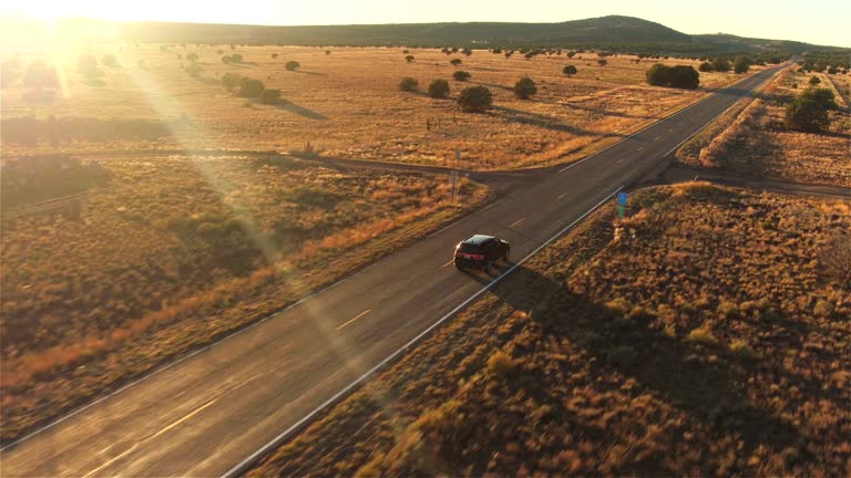 AERIAL: Black SUV car driving along empty country road at golden summer sunset