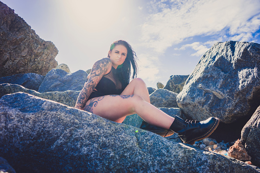 Heavily tattooed plus sized female model on South Shields beach.  She is sat on the rocks, the blue sky behind her.