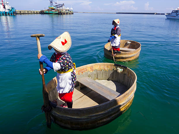 Ogi, Japan - August 19, 2014: Side view of a two woman in traditional clothes paddling the Tub-shaped wooden boats at town of Ogi n Island Sado, Sea of Japan. The boat is surrounded with sea water. In background is commercial pier with boats.