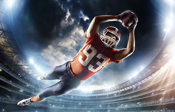 American football player jumping Wide angle low view of an American Football player jumping with a ball in the end zone for a touchdown. The action takes place on professional stadium. The player wears unbranded sports uniform. american football sport stock pictures, royalty-free photos & images