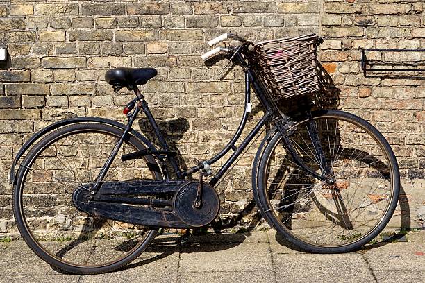 Old-Fashioned Black Bicycle With Basket Old style black ladies' bike with wicker basket leaning against a yellow brick wall in Ely, Cambridgeshire. ely england stock pictures, royalty-free photos & images