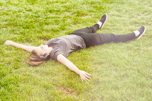 Young beautiful woman lying on grass with arms wide open and listening music on headphones on sunny day