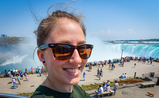 Young happy smiling woman with sunglasses on beautiful sunny springtime morning at Niagara Falls.  People & tourists in background see, hear, and feel Niagara Falls.