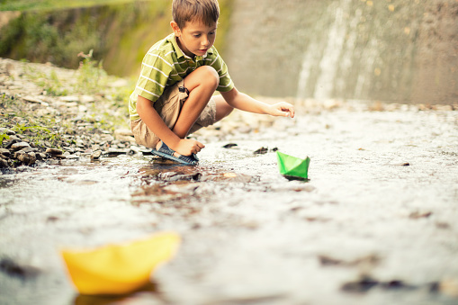 Little boy playing paper boats on the creek. He is very playful and happy while releasing ships downstream.