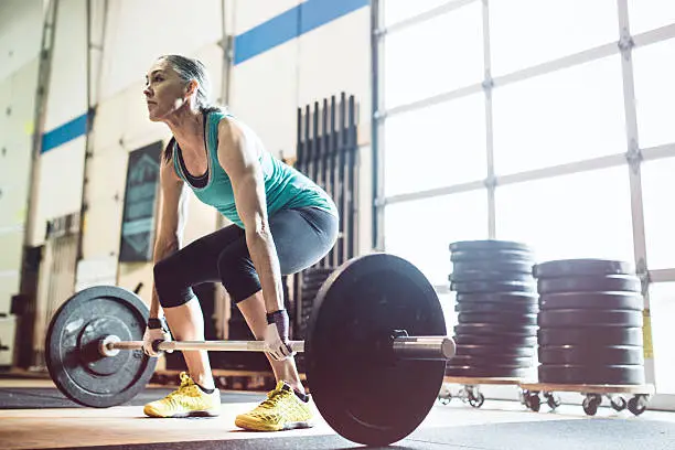 A healthy and fit woman in her 50's prepares to do an . snatch and clean in a cross training setting.  Bright clean light shines in the window behind her.  Horizontal image with copy space.