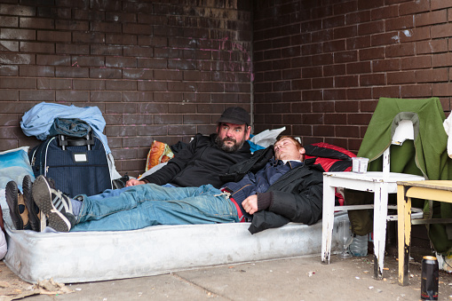 Birmingham, United Kingdom - September 4, 2015: Two homeless men lying on a matress sheltered by the overhand of a building, with rucksacs and posetions, and two white wooden traditional chairs in view. 