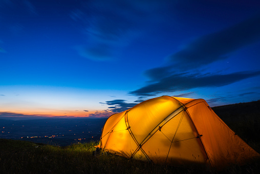 Warmly illuminated dome tent pitched on a picturesque mountain top wild camp site overlooking lights in the valley far below at sunset. ProPhoto RGB profile for maximum color fidelity and gamut.