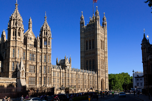 London, United Kingdom - June 30, 2015: Palace of Westminster (Houses of Parliament) as seen from Abingdon Street (Parliament Square), London, England. Victoria Tower, British flag, green trees, sightseeing tourists, bicyclists, cars and vivid blue clear sky are in the image. The image lit by evening sun.