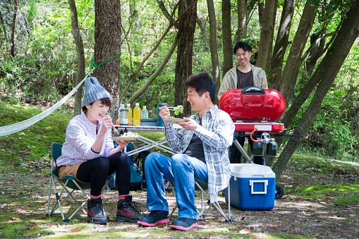 Two friends laughing and eating food, whilst a third person stands behind a BBQ at a campsite. Okayama, Japan. April 2016