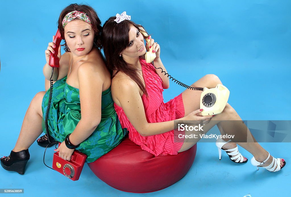 Two young women calling on phones on blue background Young woman in green dress and red phone in hand and young woman in red dress with white phone in hand calling on phone Adult Stock Photo