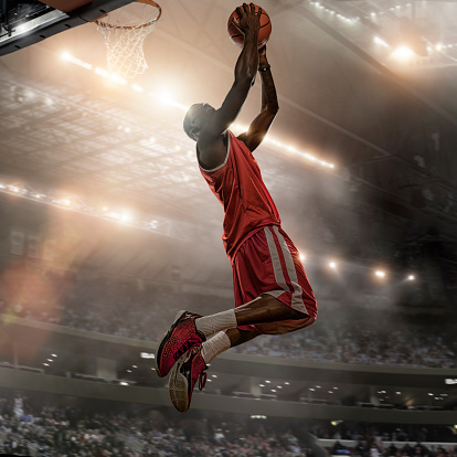 Close up image of professional basketball player holding basketball and jumping high to score a slam dunk during a basketball game. Action is set in a generic indoor basketball stadium full of spectators and with bright floodlights. With intentional light effect and lensflare. Player is dressed in generic basketball strip. 