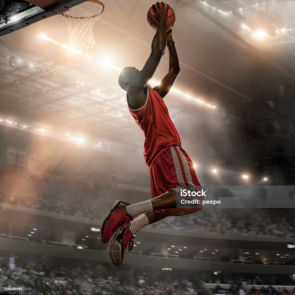 Basketball Player Action Close up image of professional basketball player holding basketball and jumping high to score a slam dunk during a basketball game. Action is set in a generic indoor basketball stadium full of spectators and with bright floodlights. With intentional light effect and lensflare. Player is dressed in generic basketball strip.  Basketball Player Stock Photo