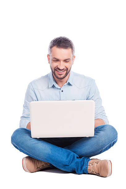 Man surfing the net. Handsome young man in casual wear sitting on the floor and working on laptop while being isolated on white background cross legged stock pictures, royalty-free photos & images
