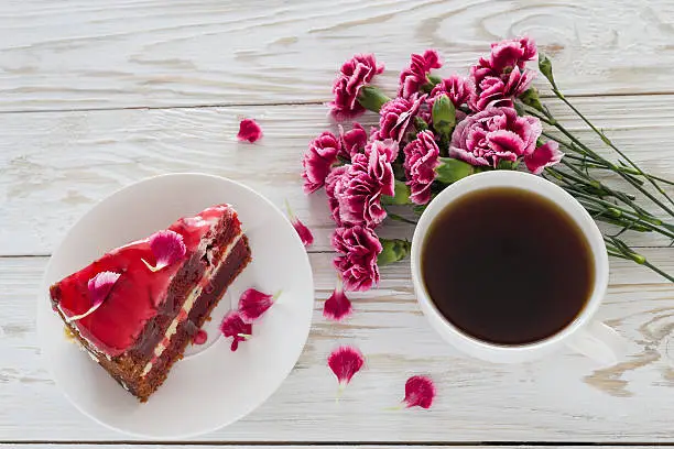 Red velvet cake, cup of coffee and pink carnations on wooden table