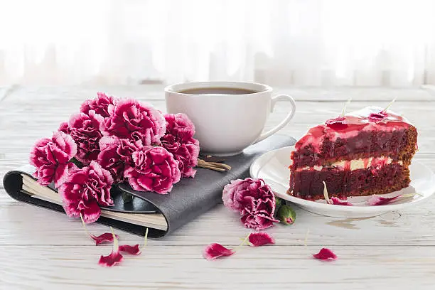 Red velvet cake, cup of coffee, notebook and pink carnations on wooden table