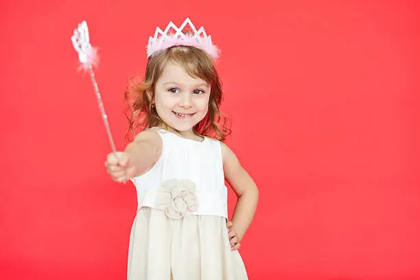Young fairy wearing white dress and crown holding a magic wand in her hand isolated over red background