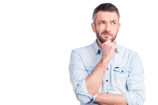 Frustrated young man in casual wear holding hand on chin and looking away while standing isolated on white background