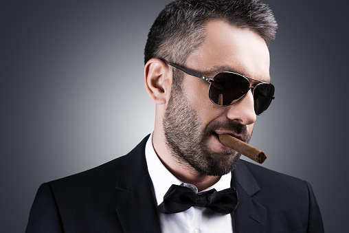 Portrait of handsome mature man in formalwear and sunglasses smoking cigar and looking away while standing against grey background
