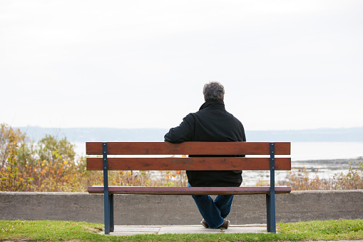 Rear view of mature man sitting on bench overlooking river.