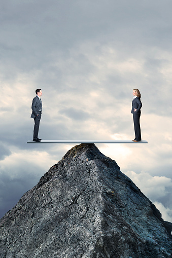 A businessman and a businesswoman  maintain their balance on a seesaw that is perched high on the pinnacle of a mountaintop. They face each otheras they maintain perfect balance and equality.