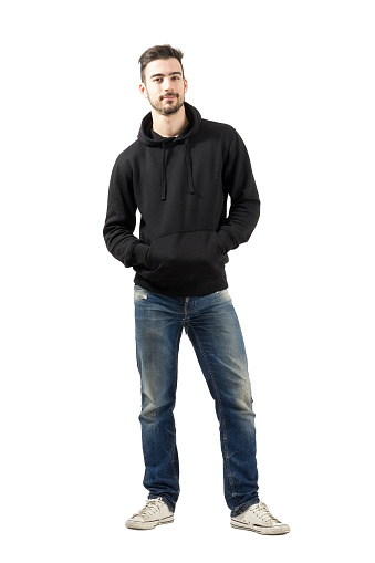 Young man in hoodie with hands in pockets smiling at camera. Full body length portrait isolated over white background.