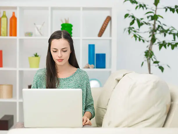 portrait of young beautiful woman sitting on sofa at home using laptop