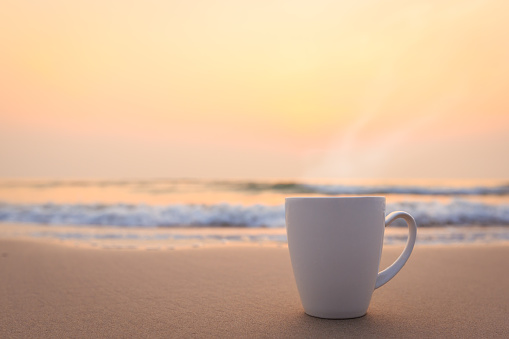 Close up white coffee cup on sand beach and view of sunset or sunrise background,peaceful