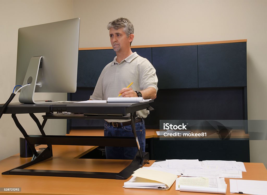 Man using stand up desk in office for good health A man is working at a standup desk in an office where he works because standing is healthier than sitting all day. Live healthy, don’t sit all day. Standing Desk Stock Photo