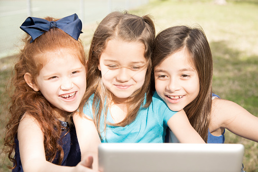 Education. Three multi-ethnic, elementary-age girl friends enjoy using a digital tablet or laptop to play educational games or video chat outside at their local park or school yard setting.  Technology.