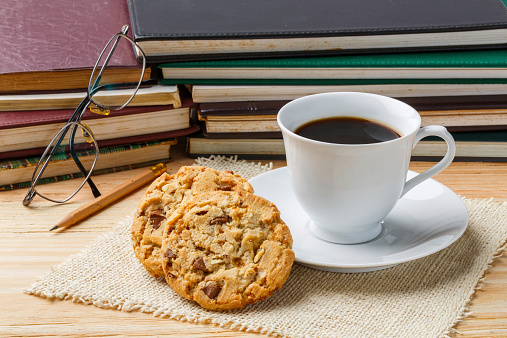 Close up dark coffee in white cup with chocolate chunk crispy cookies front of old books pencil and eyeglasses