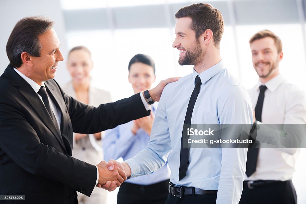 Good job! Two cheerful business men shaking hands while their colleagues applauding and smiling in the background Applauding Stock Photo