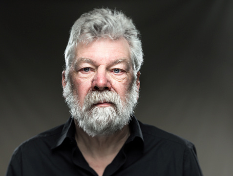 Portrait of real man, with white beard and blue eyes, looking confident straight in the camera on grey background