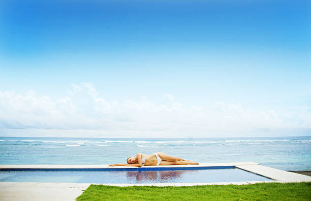istock Woman in white bikini lying by poolside against the beach 528789055