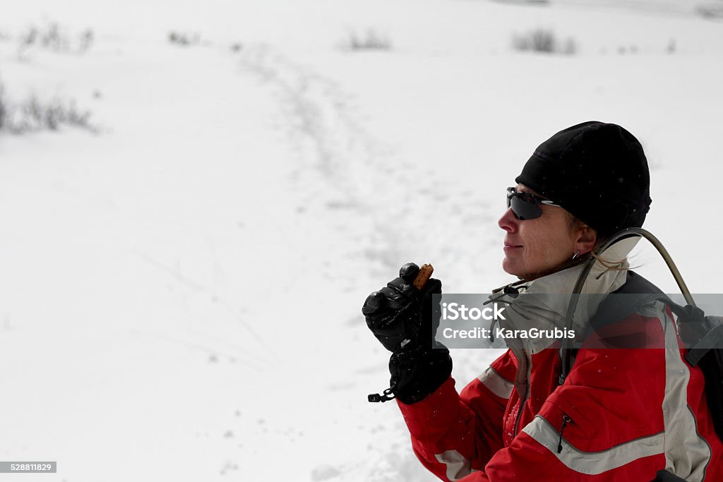 Wintertime hiker eating a nutrition bar Taken in the San Juan National Forest while snowshoeing,  Woman with sunglasses and a hydration pack stops to fuel up on a nutrition bar.  Snow and a trail are behind with ample copy space.  She appears satified with her snack. Protein Bar Stock Photo