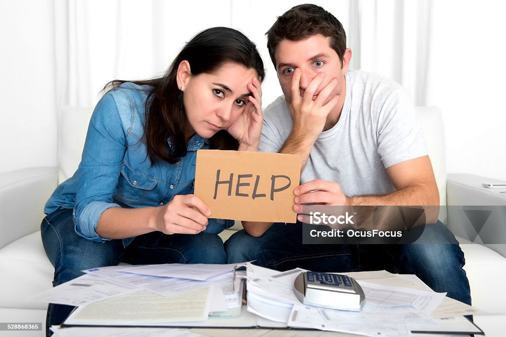 young couple in bad financial situation stress asking for help young couple worried in need of money asking for help in cardboard text message sitting in stress at home living room couch accounting debt bills bank , documents , receipts and payments feeling desperate in bad financial situation Adult Stock Photo