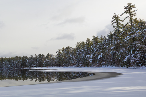 White Lake in Tamworth, NH is partially covered in ice and snow after a snow storm on a cloudy day. This lake is ringed by pine trees that are also covered in snow.