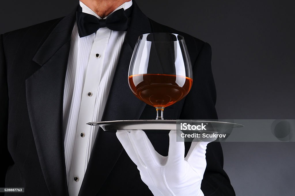 Waiter with Brandy Snifter on Tray Closeup of a waiter wearing a tuxedo and holding a tray with a brandy snifter. Low angle man is unrecognizable. Horizontal format with a light to dark gray background. Tuxedo Stock Photo