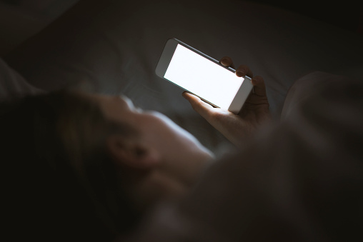 Woman using her phone under blanket in bed at night. Light of screen on her face. Blank white screen with copy space on th phone.