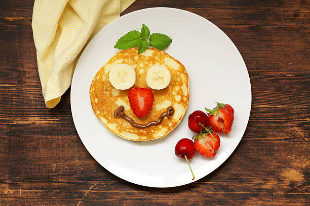 Stock photo showing close-up view of American-style, fluffy pancakes stack on white plate with split ramekin of honey and chocolate spread, lemon wedges and a banana.