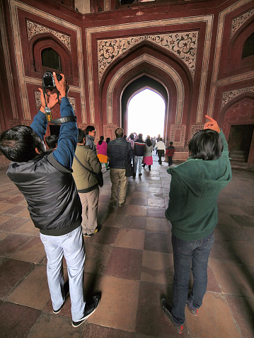 Agra, India - January 11, 2014: Back view of man and woman are taking photo of entrance to Taj Mahal from The Great gate to Taj Mahal with camera and mobile phone. In background is last gate with wall to Taj Mahal and a lot of people going trough them. It is Olympus in camera HDR.