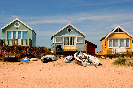 Row of three beach huts, all with blue doors. A mound of sand has blown up against them. Bright blue sky on a sunny day