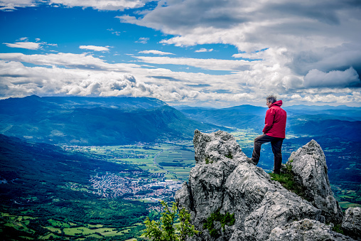 Mature man hiking in the mountains, Julian Alps, Europe. Man wearing red anorak and a rucksack is standing on a rock and looking towards Vipava valley, the city of Ajdovscina, Karst plateau and Nanos. Dramatic sky. Nikon D800, full frame, XXXL.