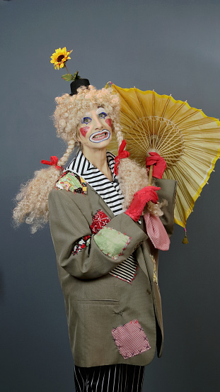 This senior female adult is dressed in a clown costume and waiting for rain. Her jacket has been extensively patched and a daisy is growing out of the derby on her wig