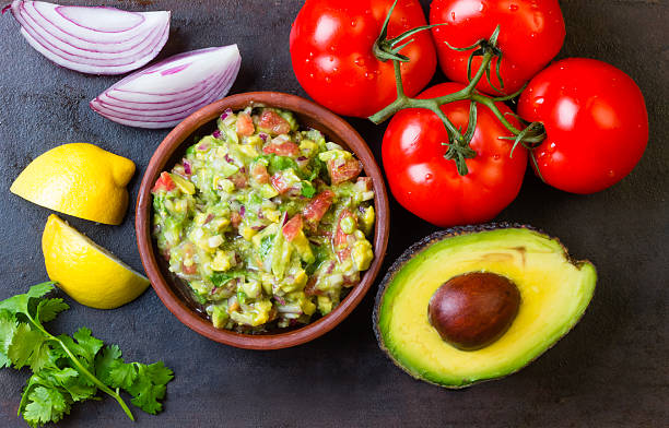 Guacamole and ingredients - avocado, tomatoes, onion, cilantro dark background. stock photo