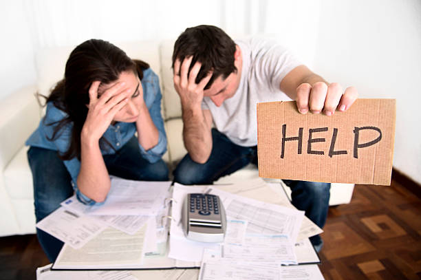 young couple in bad financial situation stress asking for help young couple worried in need of money asking for help in cardboard text message sitting in stress at home living room couch accounting debt bills bank , documents , receipts and payments feeling desperate in bad financial situation financial needs stock pictures, royalty-free photos & images