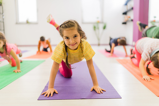 Little girl doing stretching exercises with her leg up and looking at the camera, while her friends are around her.