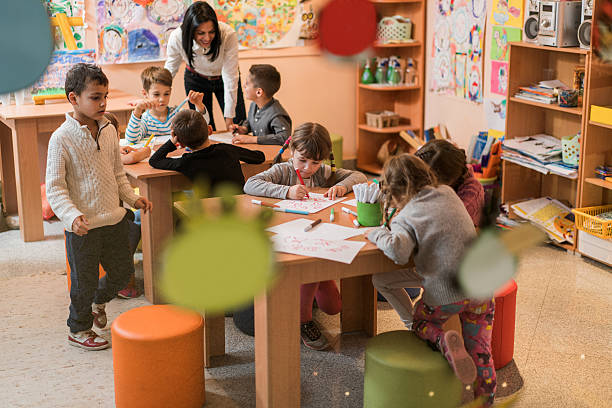 Large group of children and their teacher in a classroom at preschool. Focus is on girl with ponytails drawing. The view is through glass.