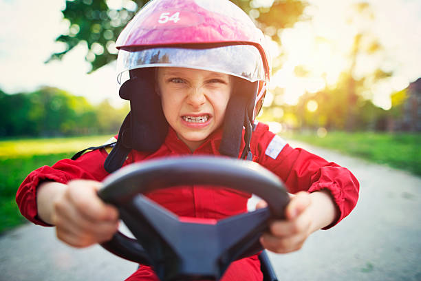 Portrait of furious little boy riding a fast go-kart. Little boy aged 6 driving a pedal go-kart, kart, soapbox car or cyclekart. Little boy is angry and clenching his teeth. He is obviously very focused on winning the race. Road and sunset visible in the background. soapbox cart stock pictures, royalty-free photos & images